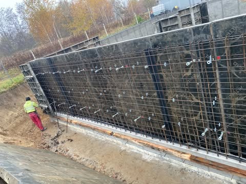 Przygotowanie szalunku i zbrojenia pod wylewkę fundamentów domu jednorodzinnego z podpiwniczeniem, widoczne metalowe elementy mocujące i pracownik w odzieży roboczej.