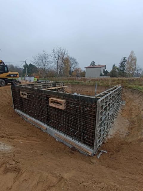Szare szalunki zbrojeniowe w wykopie pod budowę domu jednorodzinnego z podpiwniczeniem, widoczny żółty dźwig w tle i pochmurne niebo.