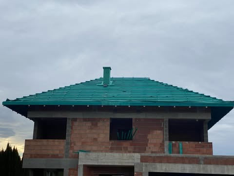 Wznoszony dom jednorodzinny z cegły, z zielonym dachem ceramicznym i kominem, stan surowy otwarty, widoczne zbrojenia.