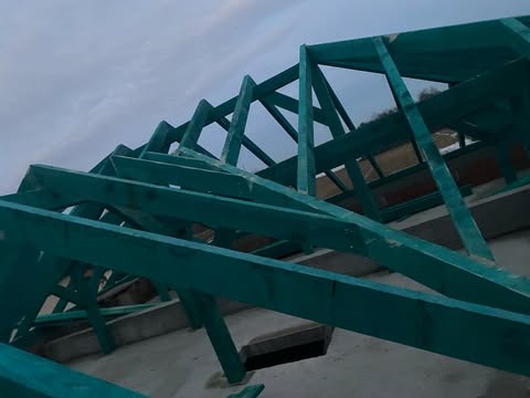 Drewniana konstrukcja więźby dachowej, pomalowana na zielono, widoczna z dołu na tle pochmurnego nieba.