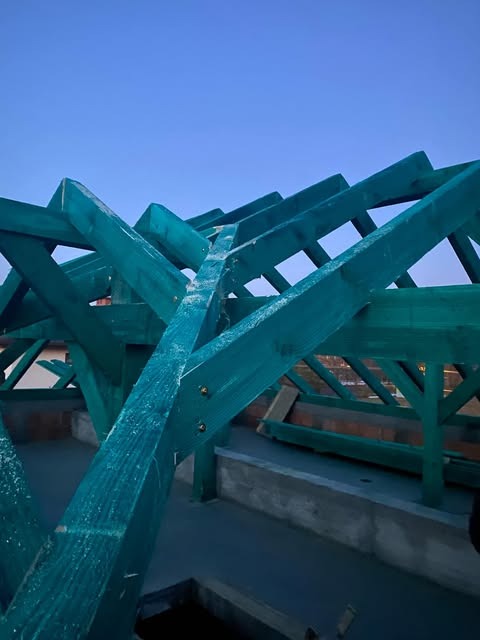 Konstrukcja drewniana dachu, pomalowana na zielono, widoczna od spodu na tle błękitnego nieba. Elementy połączone śrubami. Widoczna izolacja fundamentów.