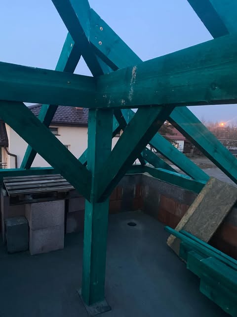 Wiązary dachowe z impregnowanego drewna w kolorze zielonym, widok z góry na budowę domu jednorodzinnego, w tle budynek mieszkalny z ciemną dachówką.