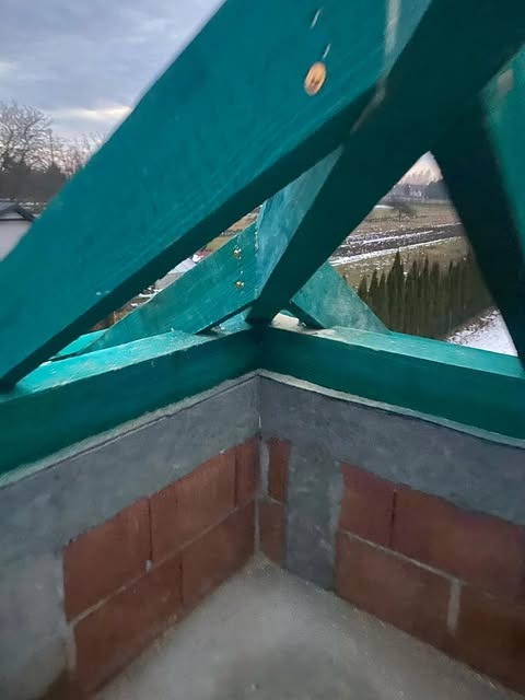 Wewnętrzny narożnik ściany z czerwonej cegły, częściowo otynkowanej, z widoczną konstrukcją drewnianą dachu pomalowaną na zielono, widok na zewnątrz na drzewa i pole.
