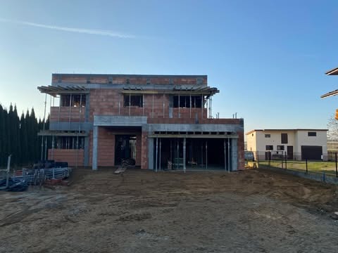 Dwukondygnacyjny dom jednorodzinny w trakcie budowy z cegły i betonu, widoczne szalunki na stropie i podpory, teren wokół w stanie surowym, w tle zabudowa jednorodzinna.