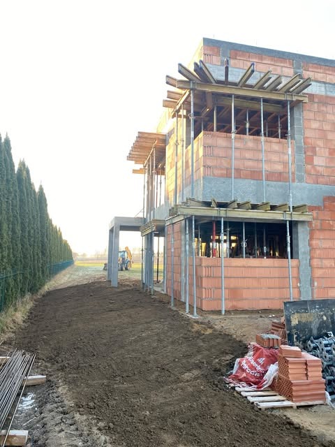 Wznoszenie dwukondygnacyjnego domu murowanego z czerwonej cegły, widoczne szalunki na oknach, teren wokół wyrównany, w tle koparka.