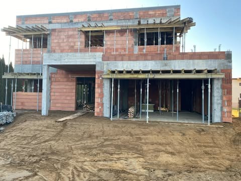 Wznoszenie dwukondygnacyjnego domu jednorodzinnego z czerwonej cegły, widoczne szalunki stropowe i podpory, teren wokół w stanie surowym.