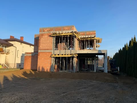 Budowa domu jednorodzinnego w Tarnowie w stanie surowym otwartym, widoczne ściany z czerwonej cegły, szalunki i elementy zbrojeniowe, na działce stoi mała koparka.