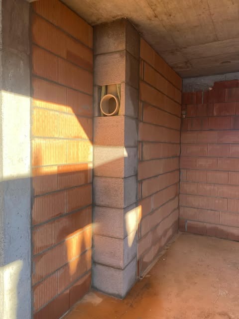 Wnętrze w trakcie budowy domu jednorodzinnego: ściany z czerwonej cegły, betonowy filar, pionowy kanał wentylacyjny z rurą z tworzywa w narożniku.