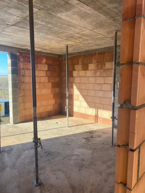 Wnętrze budowanego domu jednorodzinnego z czerwonej cegły, ściany w trakcie wznoszenia, betonowy strop podparty metalowymi stemplami budowlanymi, widok z zewnątrz przez otwór okienny.