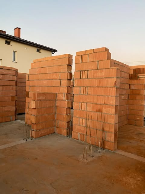 Wznoszenie ścian parteru z czerwonej ceramiki poryzowanej na budowie domu jednorodzinnego, wystające pręty zbrojeniowe, widok na sąsiedni budynek.