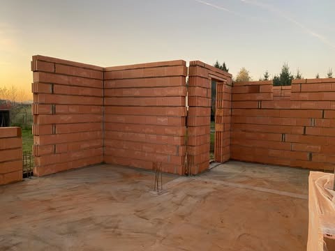 Wznoszone mury domu jednorodzinnego z czerwonej cegły ceramicznej Porotherm, widoczne fragmenty zbrojenia, stan surowy otwarty, tło stanowią drzewa i zachodzące słońce.