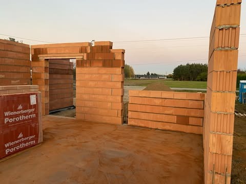 Wznoszenie ścian parteru domu jednorodzinnego z bloczków ceramicznych Porotherm, widoczne fragmenty ścian, otwór drzwiowy, paleta bloczków, w tle hałda ziemi i zabudowania.