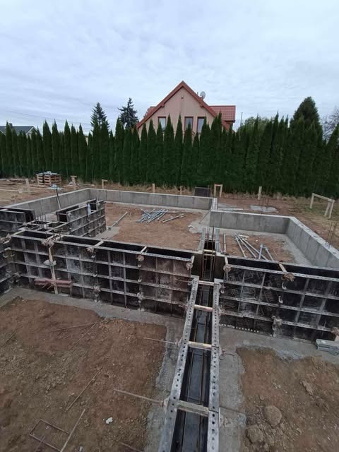 Przygotowanie pod fundamenty domu jednorodzinnego: szalunki ustawione na obrysie, widoczne zbrojenia i wykop, w tle rząd wysokich tuj oraz dom z czerwonym dachem.