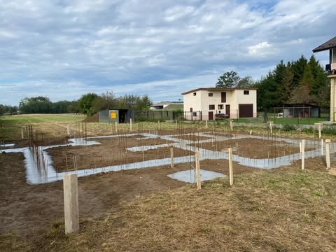 Fundamenty domu jednorodzinnego w Bogumiłowicach: zalane betonem ławy fundamentowe z wystającymi prętami zbrojeniowymi, widok na budynek gospodarczy i dom w tle.