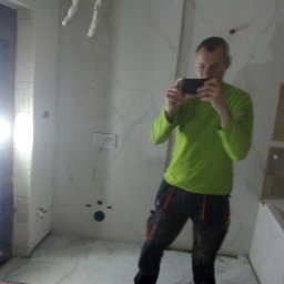 Przemysław Rudnicki - Mężczyzna w zielonej koszulce robi selfie w trakcie prac wykończeniowych w łazience z marmurowymi płytami, widoczne przygotowane otwory na armaturę i częściowo zamaskowane rury.