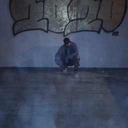 Przemysław Rudnicki - Mężczyzna w czapce i rękawicach roboczych kuca na podłodze w pomieszczeniu z graffiti na ścianie.