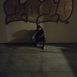 Przemysław Rudnicki - Mężczyzna w czapce i kraciastej koszuli kuca na tle ściany z graffiti w słabym oświetleniu.