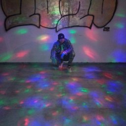 Przemysław Rudnicki - Mężczyzna w czapce i flanelowej koszuli kuca na podłodze w pokoju z graffiti na ścianie i kolorowymi światłami dyskotekowymi.