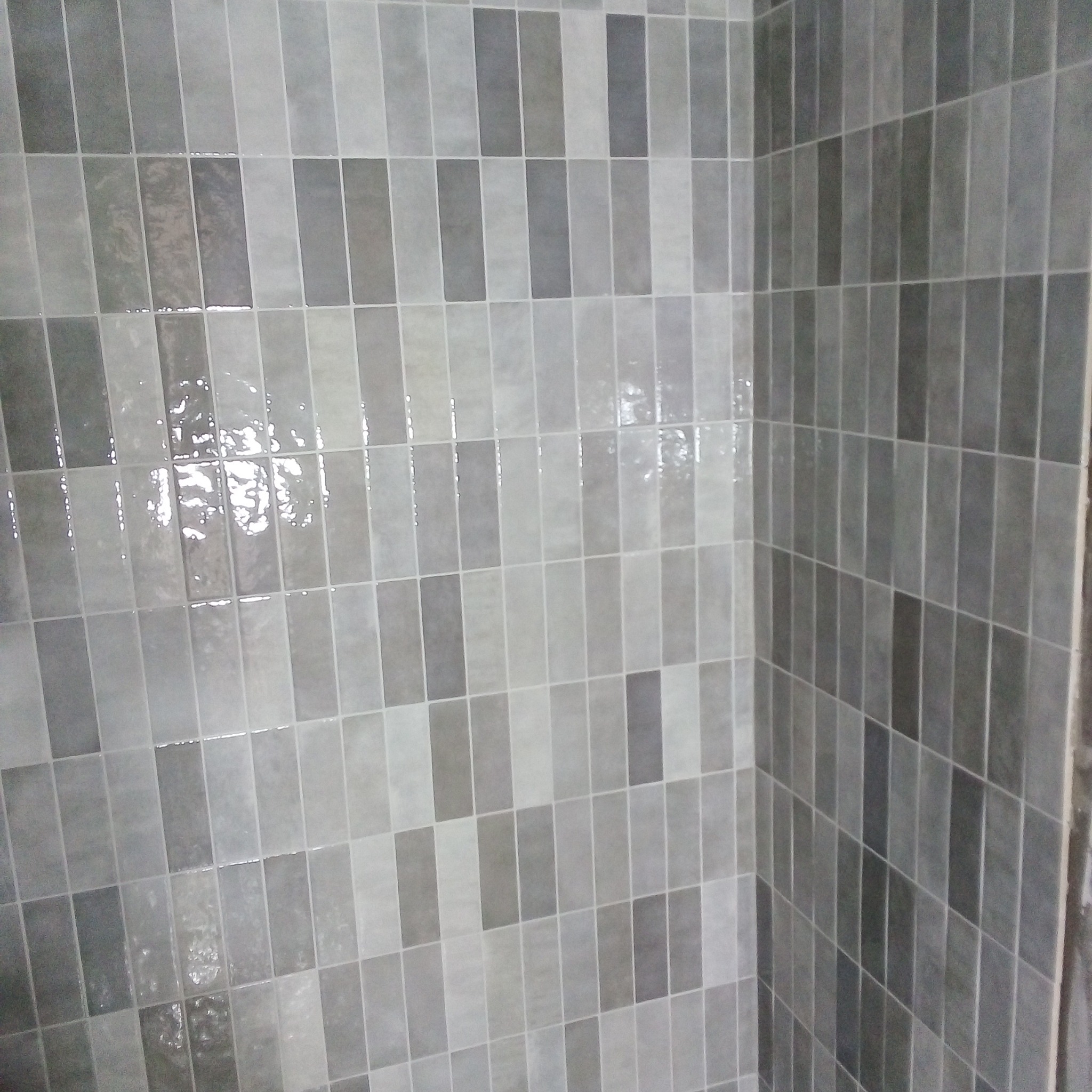 Ściana wyłożona błyszczącymi, prostokątnymi płytkami w odcieniach szarości, ułożonymi w pionowy wzór, widoczny narożnik pomieszczenia.