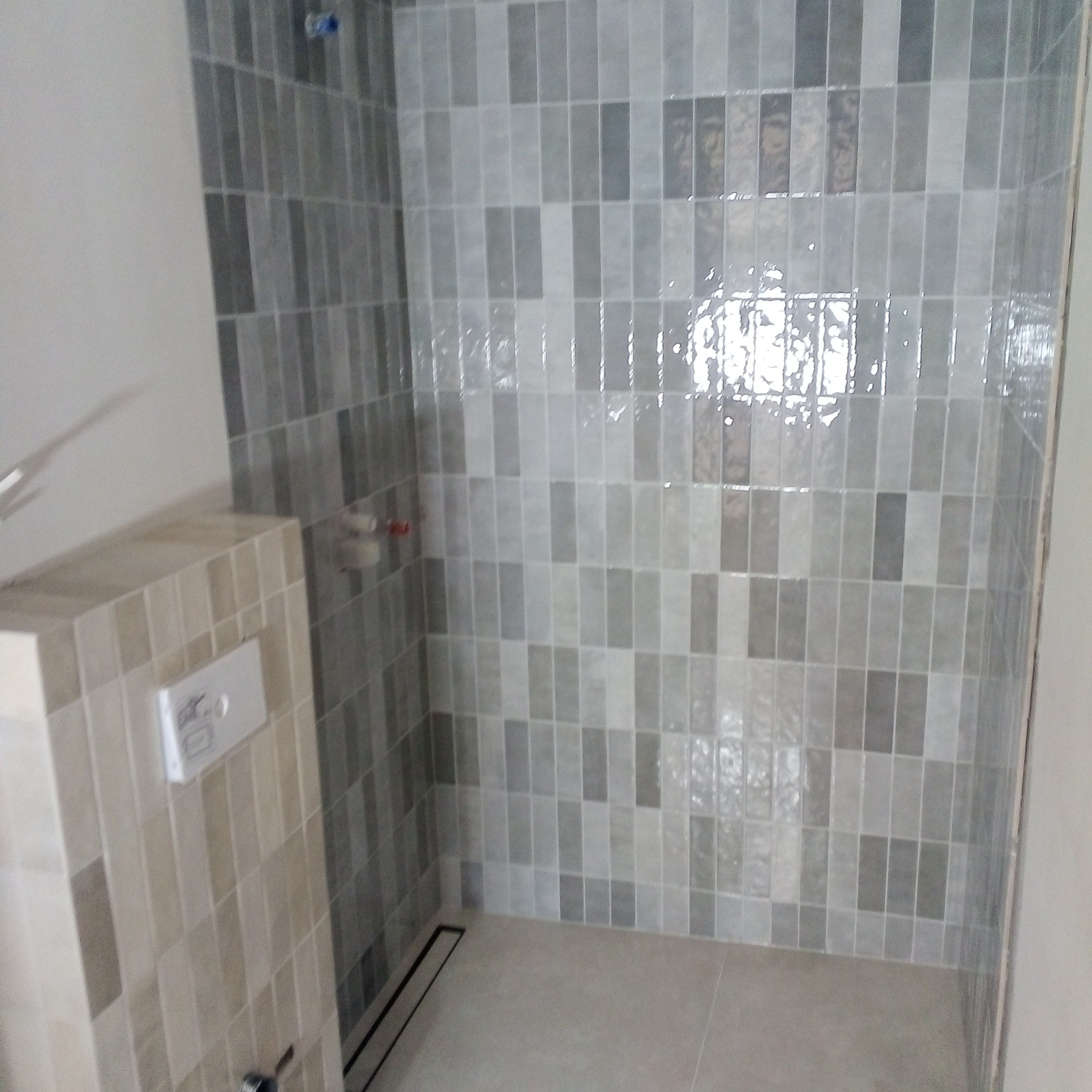 Wykończona kabina prysznicowa z szarymi, podłużnymi płytkami ułożonymi pionowo, widoczny odpływ liniowy i fragment zabudowy WC z beżowych płytek.