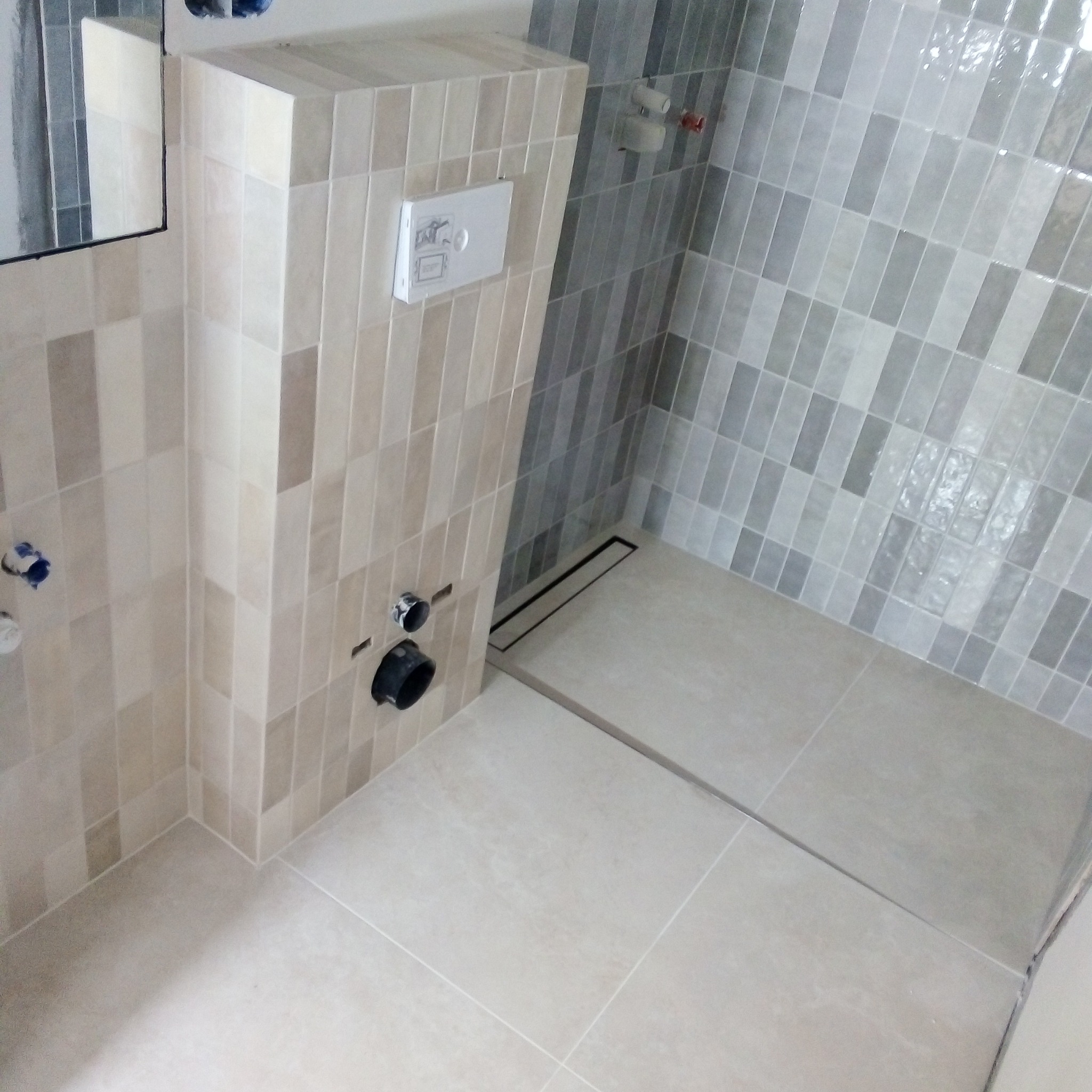 Wykończona łazienka z beżowymi i szarymi płytkami w pionowy wzór, zabudowa stelaża podtynkowego WC z przyciskiem spłukującym, odpływ liniowy w podłodze.