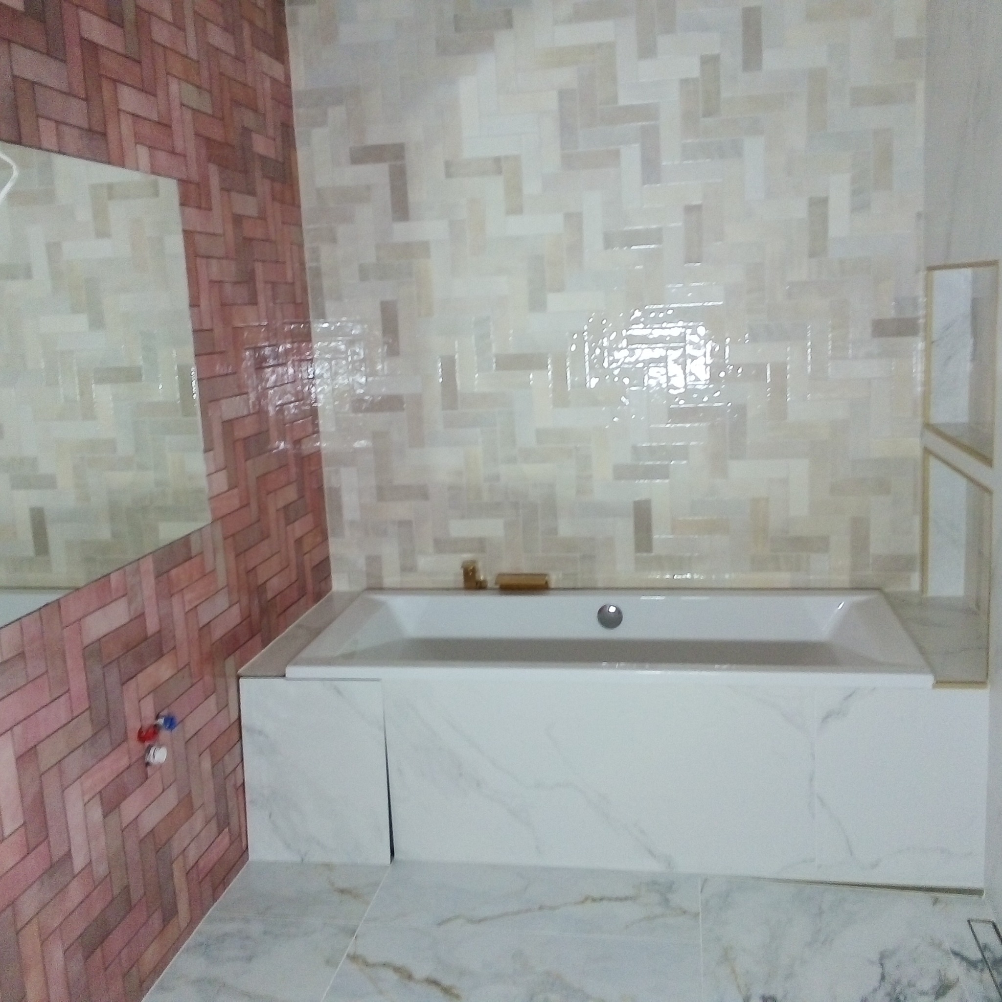 Wykończona łazienka z wanną obudowaną białymi płytami imitującymi marmur, ściany wyłożone kafelkami w odcieniach beżu i brązu ułożonymi we wzór jodełki, widoczne elementy instalacji wodnej.