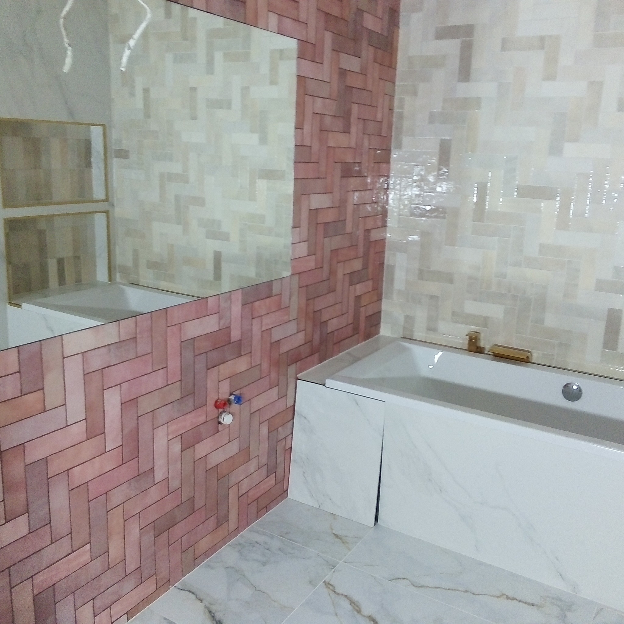 Wykończona łazienka z wanną obudowaną marmurowymi płytami, ściany wyłożone płytkami w odcieniach różu i beżu ułożonymi w jodełkę, lustro odbijające fragment łazienki.