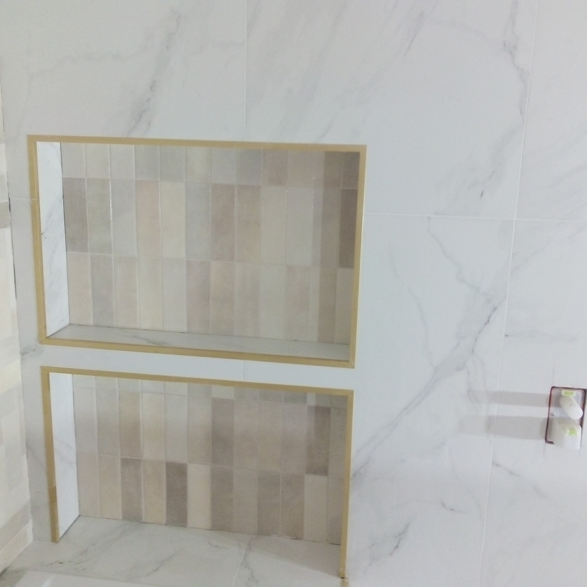 Dwie płytkie, prostokątne wnęki wyłożone pionowymi płytkami w odcieniach beżu i szarości, obramowane złotą listwą, wkomponowane w ścianę z białych płytek imitujących marmur.