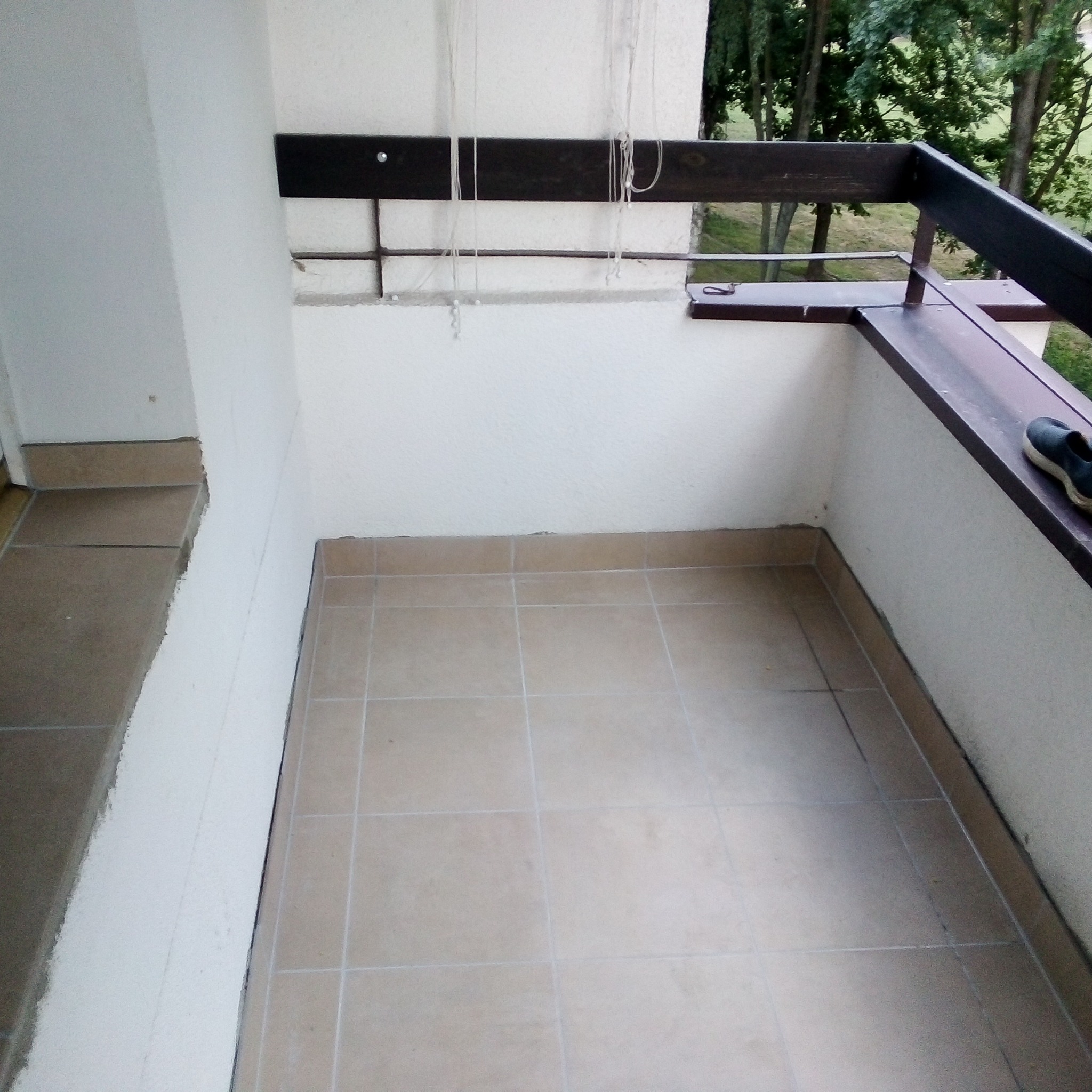 Jasne płytki ceramiczne na balkonie z drewnianą balustradą i widokiem na drzewa w tle, jedno niebieskie obuwie leży na balustradzie.