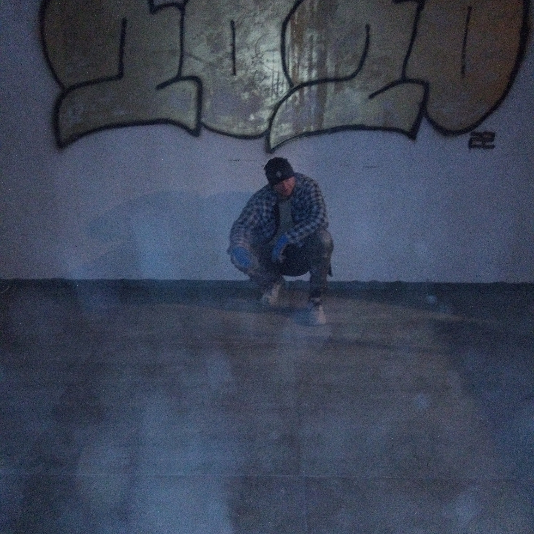 Mężczyzna w czapce i rękawicach roboczych kuca na podłodze w pomieszczeniu z graffiti na ścianie.