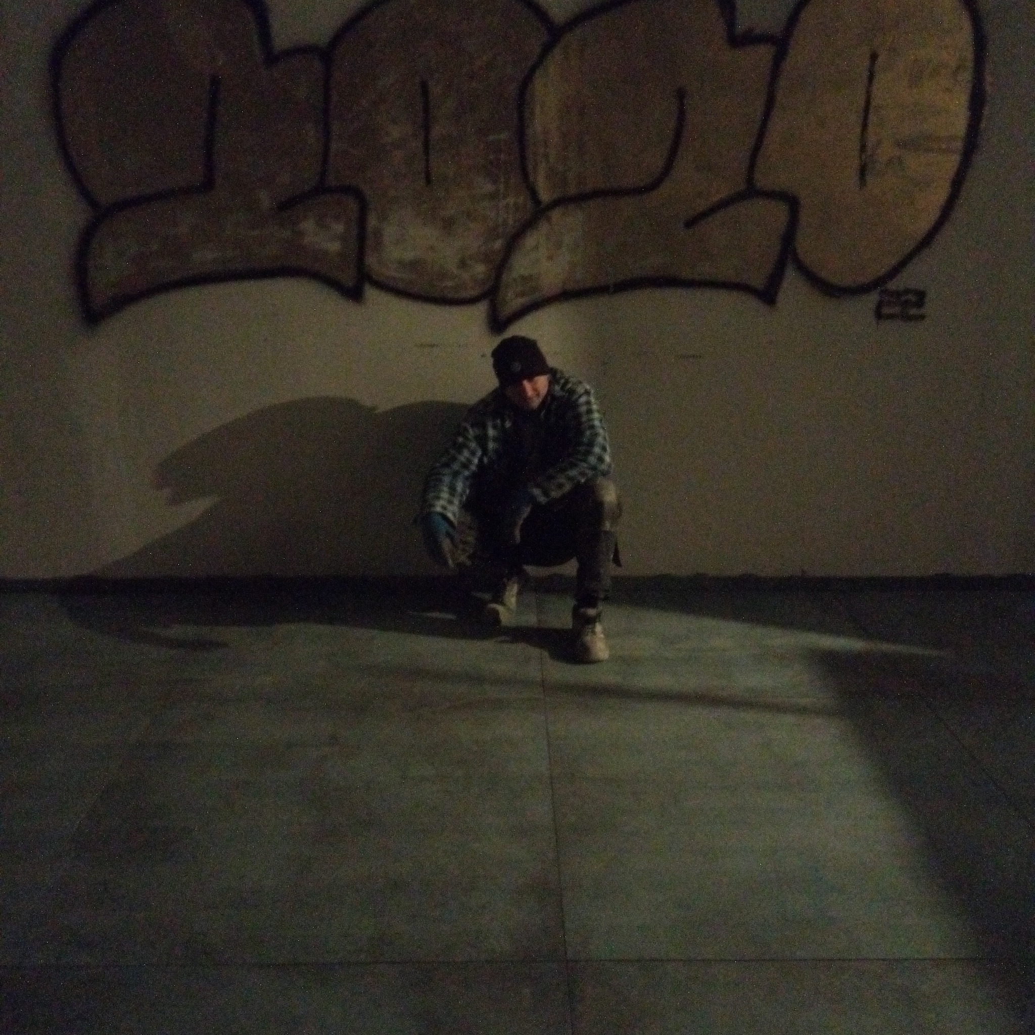 Mężczyzna w czapce i kraciastej koszuli kuca na tle ściany z graffiti w słabym oświetleniu.