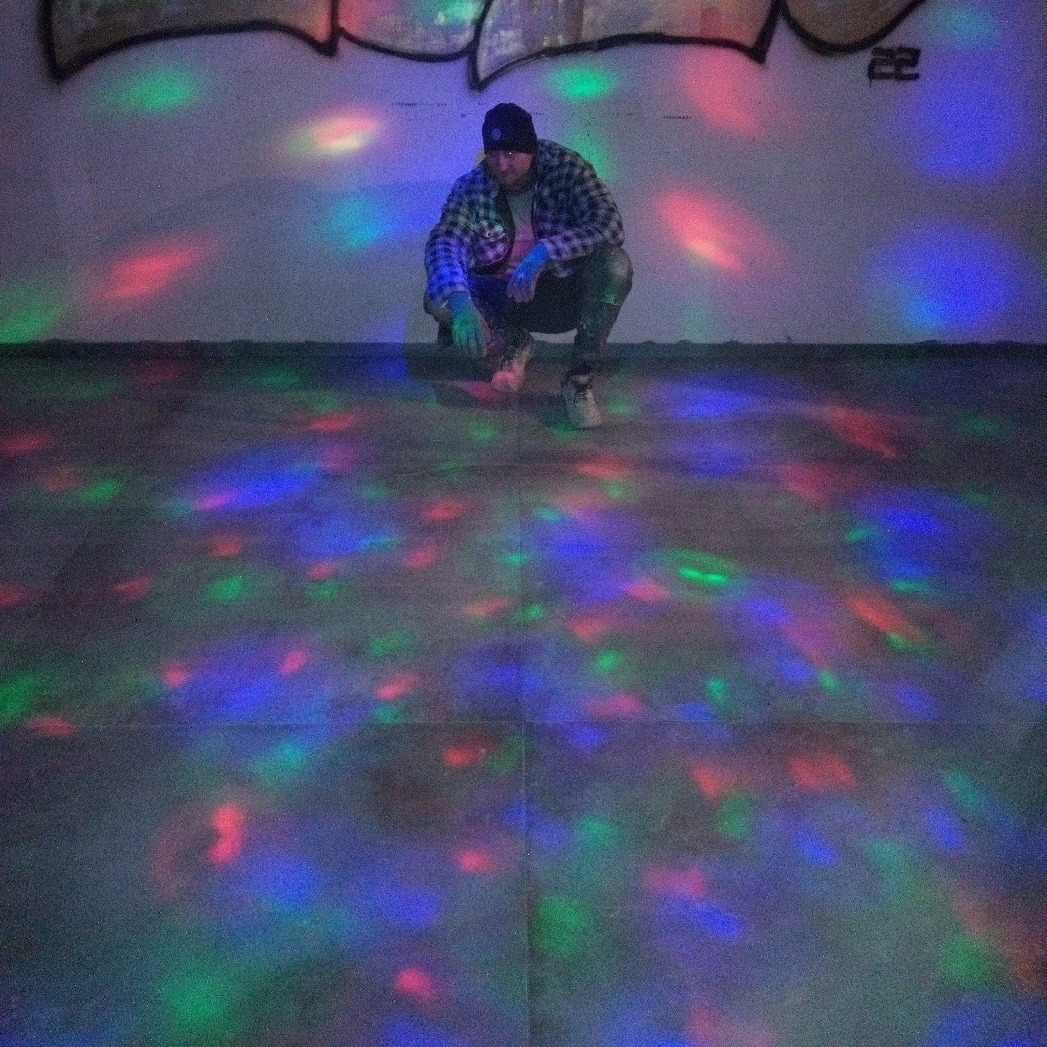 Mężczyzna w czapce i flanelowej koszuli kuca na podłodze w pomieszczeniu z graffiti i kolorowymi światłami dyskotekowymi.