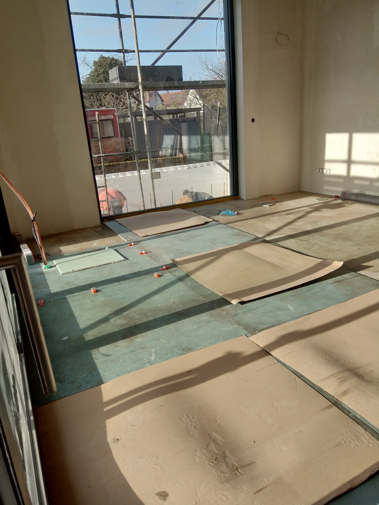 Przygotowanie podłoża pod nową podłogę: wyrównana wylewka, zabezpieczona kartonem, widoczne elementy poziomujące, duże okno z widokiem na rusztowania i okoliczne budynki.
