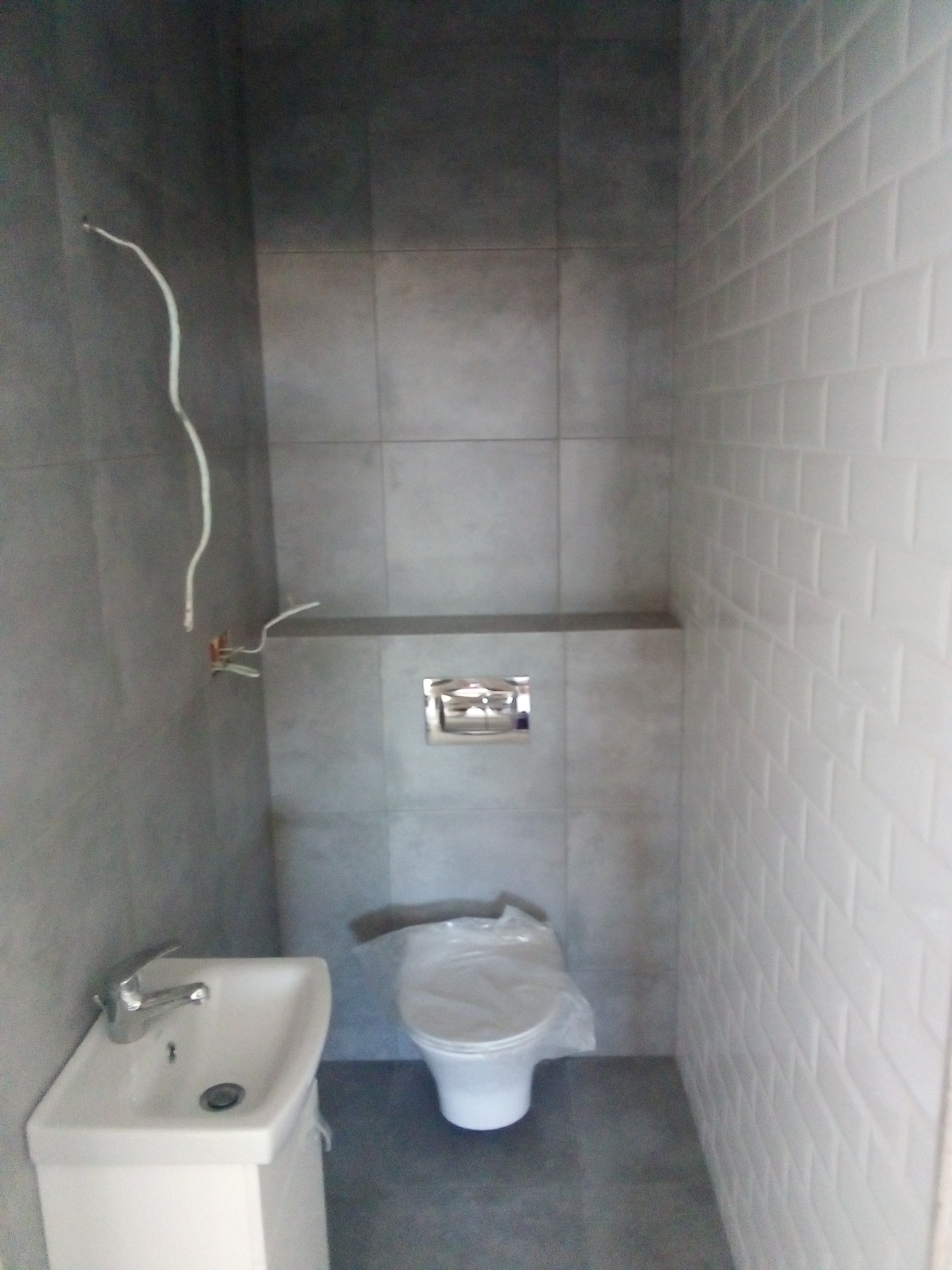 Wnętrze małej łazienki w trakcie remontu, widoczne szare płytki na ścianach, białe płytki typu metro, biała umywalka z baterią, toaleta przykryta folią ochronną oraz wystające przewody elektryczne.