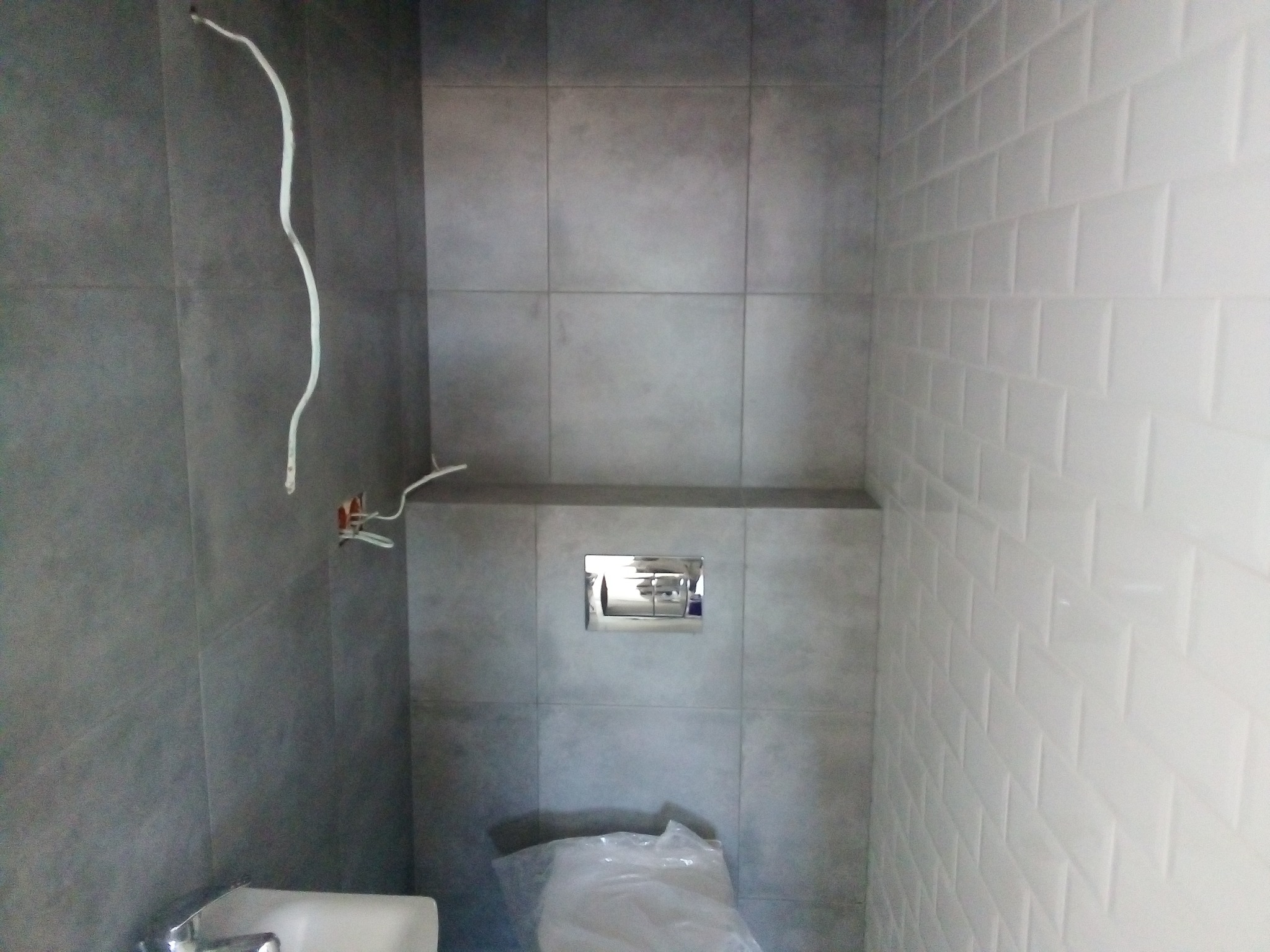 Wnętrze łazienki w trakcie prac wykończeniowych: szare płytki wielkoformatowe na jednej ścianie, białe kafelki typu 'metro' na drugiej, wystające przewody elektryczne i rury hydrauliczne, toaleta...