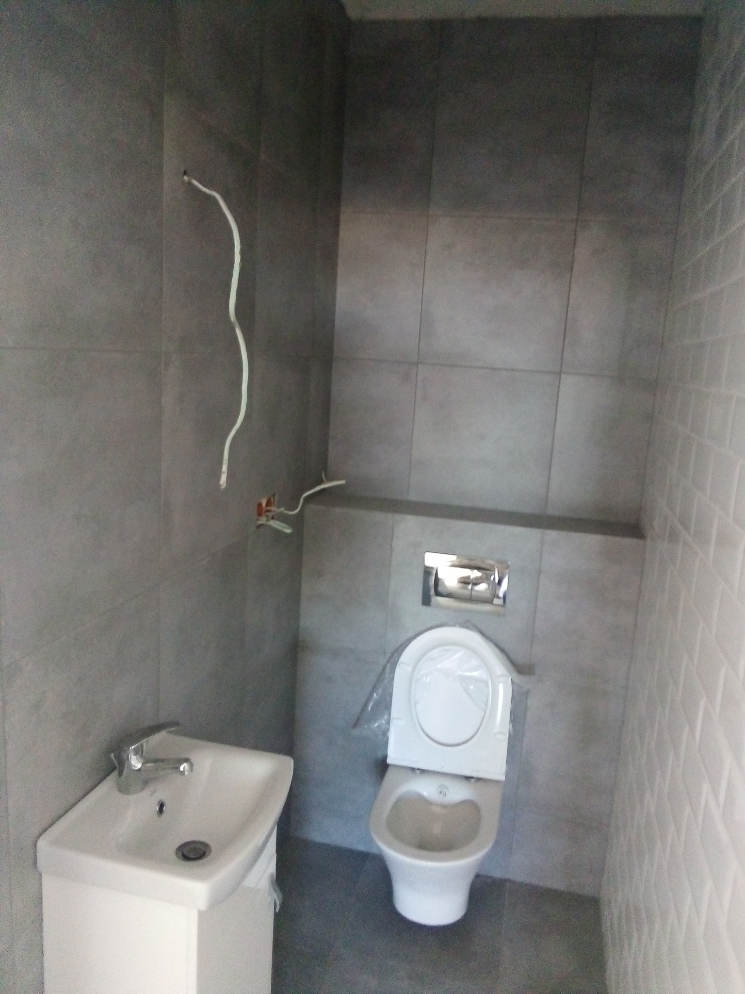 Szara łazienka w trakcie wykańczania: widoczne kable wystające ze ściany, biała umywalka z szafką i toaleta z plastikową osłoną na desce, płytki w dwóch odcieniach szarości.