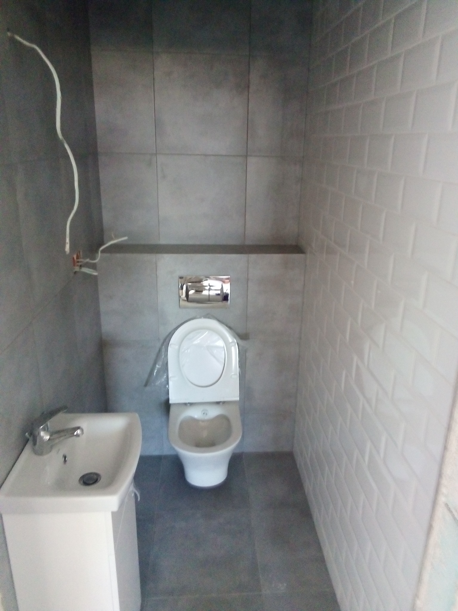 Nowa łazienka w szaro-białej kolorystyce z umywalką na szafce, wiszącą toaletą z chromowanym przyciskiem spłukiwania i częściowo wykończonymi instalacjami elektrycznymi.