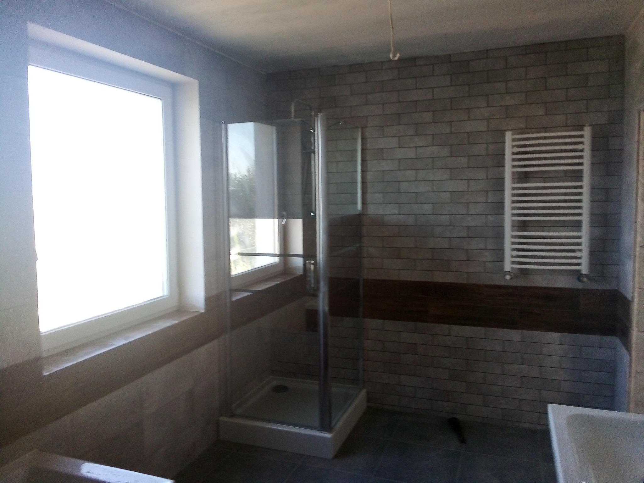 Szara łazienka po remoncie z kabiną prysznicową narożną, grzejnikiem drabinkowym i oknem z widokiem na zieleń, wykończona płytkami imitującymi cegłę.