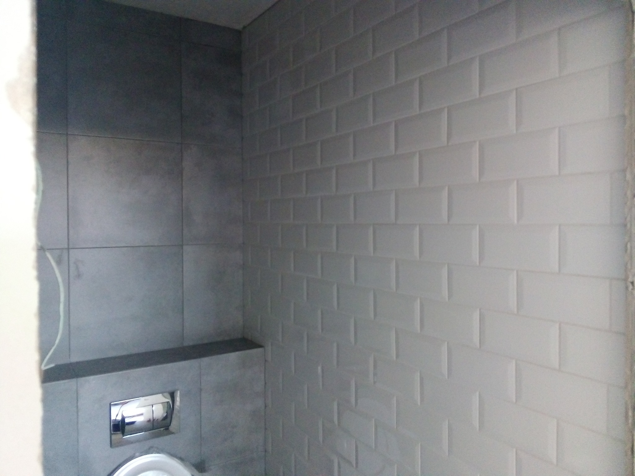 Ściana łazienki wyłożona białymi, prostokątnymi płytkami typu metro, kontrastująca z szarymi płytami i zabudową stelaża podtynkowego toalety.