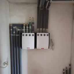 Dwa białe rozdzielacze ogrzewania podłogowego marki Onnline, zamontowane na ścianie w surowym stanie, połączone izolowanymi rurami grzewczymi. Widoczne fragmenty instalacji podłogowej Reflex...