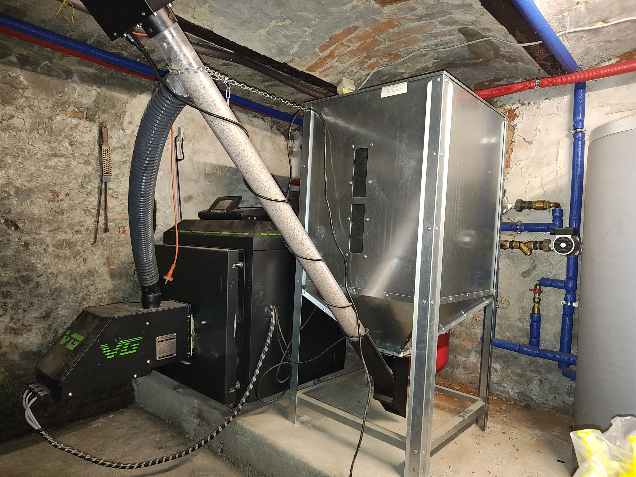 Nowoczesny piec na pellet z podajnikiem w kotłowni o surowych ścianach, widoczne rury instalacji grzewczej i zbiornik na wodę.
