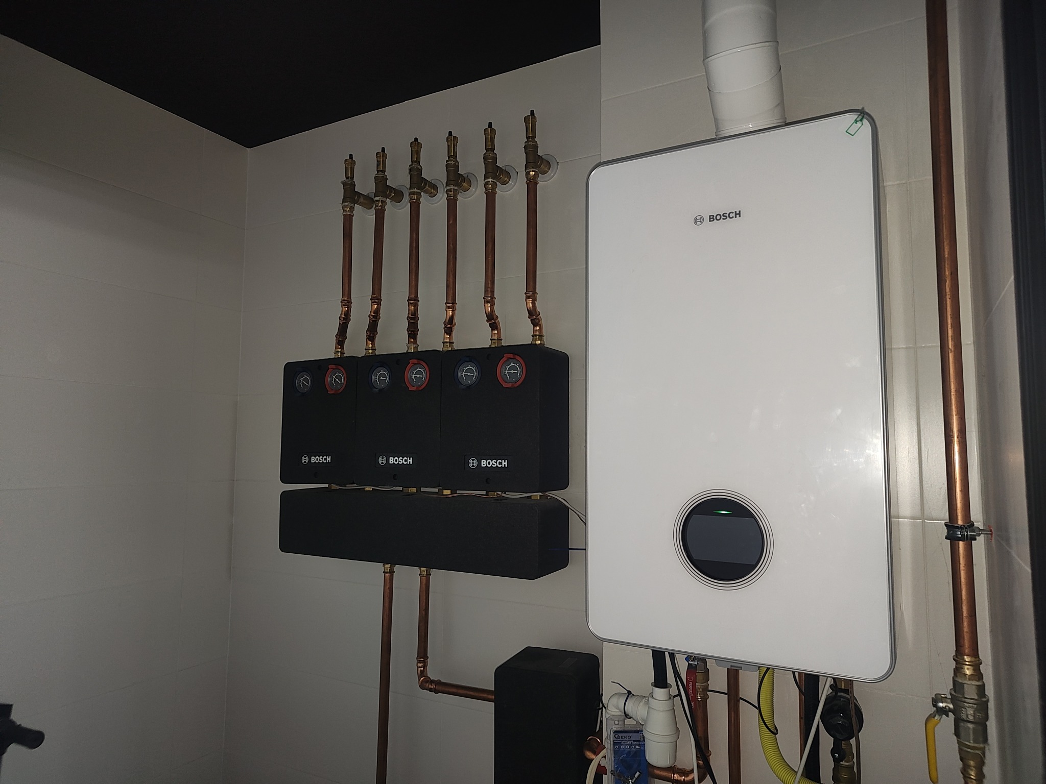 Nowoczesny, biały piec gazowy marki Bosch z czarnymi elementami sterującymi i miedzianymi rurami w pomieszczeniu z białymi płytkami i czarnym sufitem.