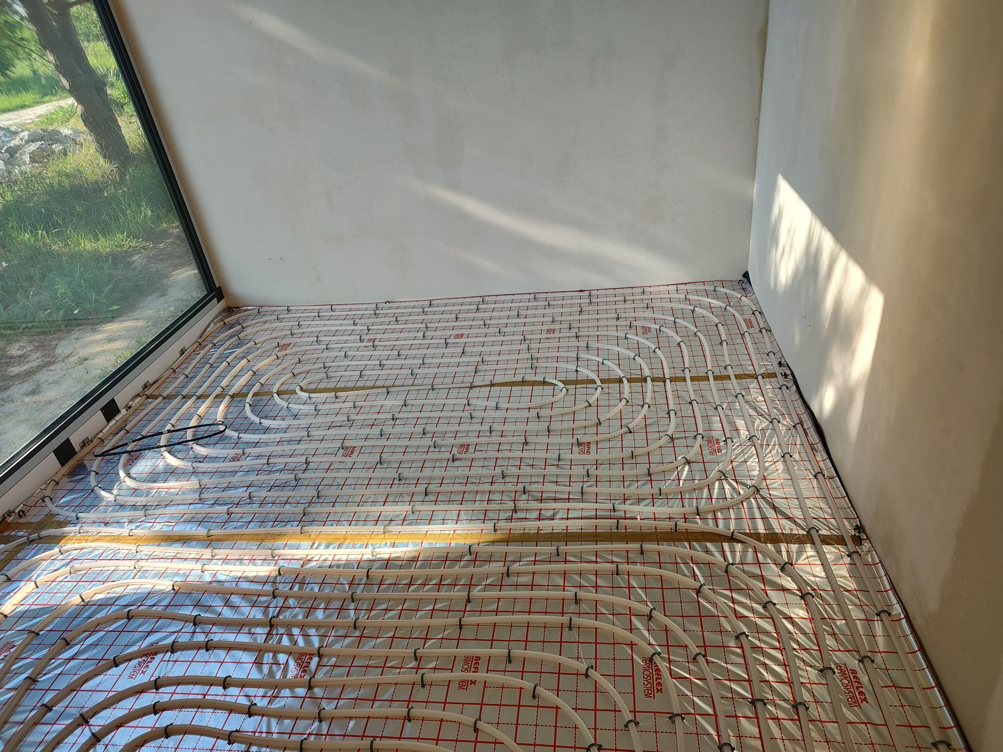 Białe rury ogrzewania podłogowego ułożone w pętle na folii z czerwoną siatką, widoczne przy oknie i surowej ścianie w nowym budownictwie.