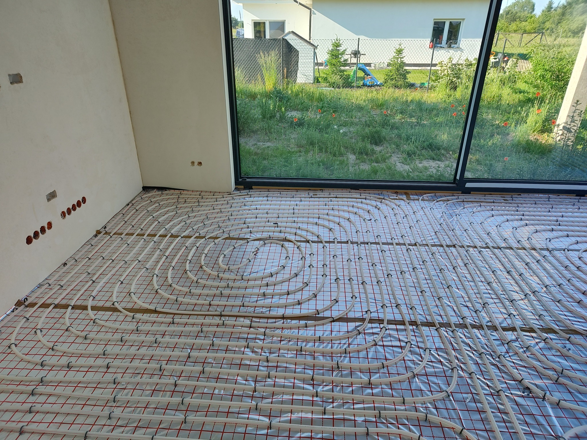 Rozłożona instalacja ogrzewania podłogowego na folii izolacyjnej z widocznymi rurami w kształcie spirali, w tle okno z widokiem na ogród.