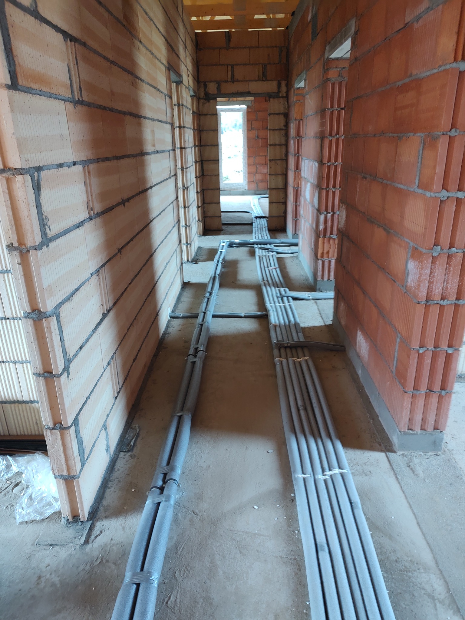 Wnętrze budynku w trakcie budowy z widocznymi przewodami instalacji grzewczej prowadzonymi po podłodze, zabezpieczonymi szarą izolacją i taśmą, ściany z czerwonej cegły kratówki.