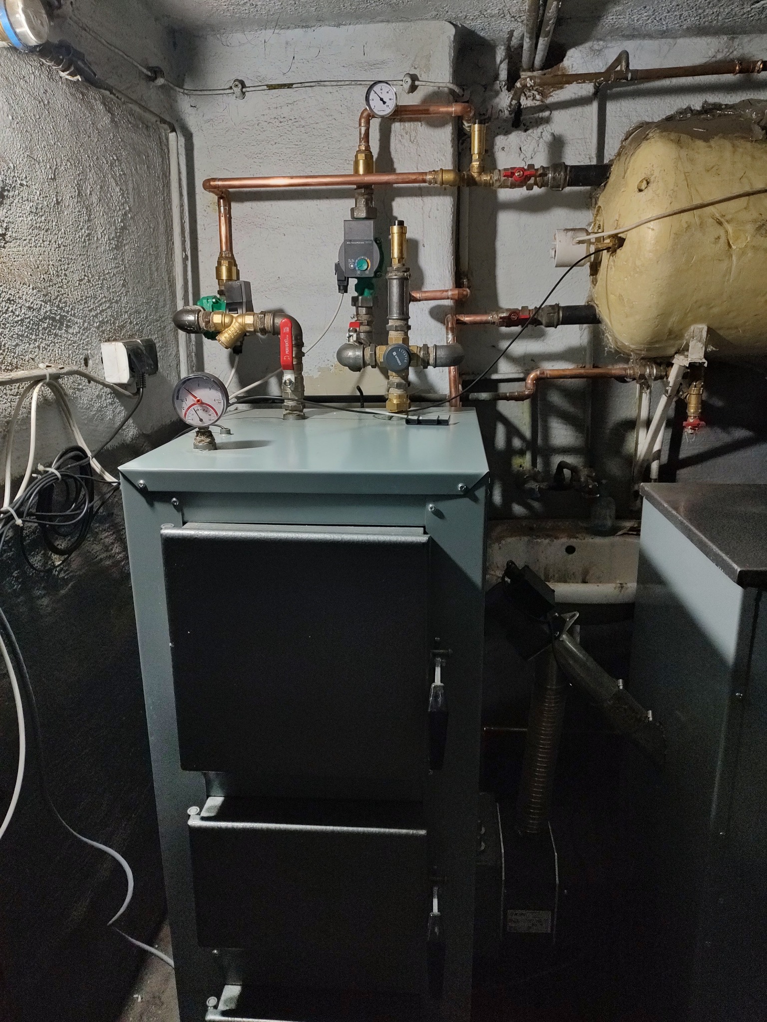 Nowoczesny piec centralnego ogrzewania z manometrem, podłączony do instalacji z miedzianych rur i zaworów w piwnicy z szorstką, pomalowaną ścianą.