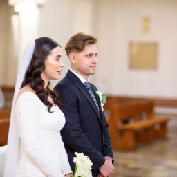 NIKA KHRYSTYNA NEDOMOLKINA - Para młoda w kościele podczas ceremonii ślubnej. Panna młoda w białej sukni z welonem, pan młody w granatowym garniturze. W tle ławki i drewniane drzwi.