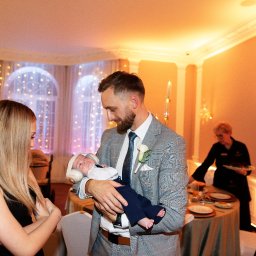 NIKA KHRYSTYNA NEDOMOLKINA - Elegancki mężczyzna w garniturze trzyma śpiące niemowlę w słuchawkach na tle sali bankietowej z dekoracjami świetlnymi i gośćmi. Ujęcie z uroczystości rodzinnej.