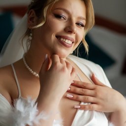 NIKA KHRYSTYNA NEDOMOLKINA - Uśmiechnięta panna młoda w białym szlafroku z piórami, perłowym naszyjnikiem i diademem, z delikatnym makijażem, pozuje do portretu w jasnym wnętrzu.