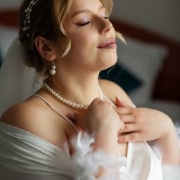 NIKA KHRYSTYNA NEDOMOLKINA - Portret panny młodej w satynowym szlafroku z piórami, z perłami i tiarą, w delikatnym makijażu ślubnym, uchwycony w miękkim świetle w Poznaniu. Zamknięte oczy, spokój.